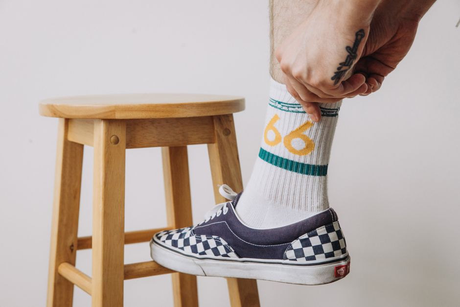 A tattooed hand adjusts a sock above a checkered shoe on a wooden stool, highlighting casu