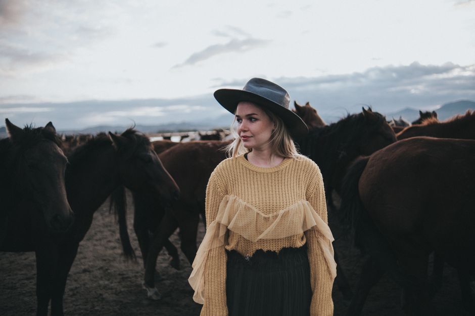 Woman in a stylish outfit and hat standing among a herd of horses outdoors at dusk.