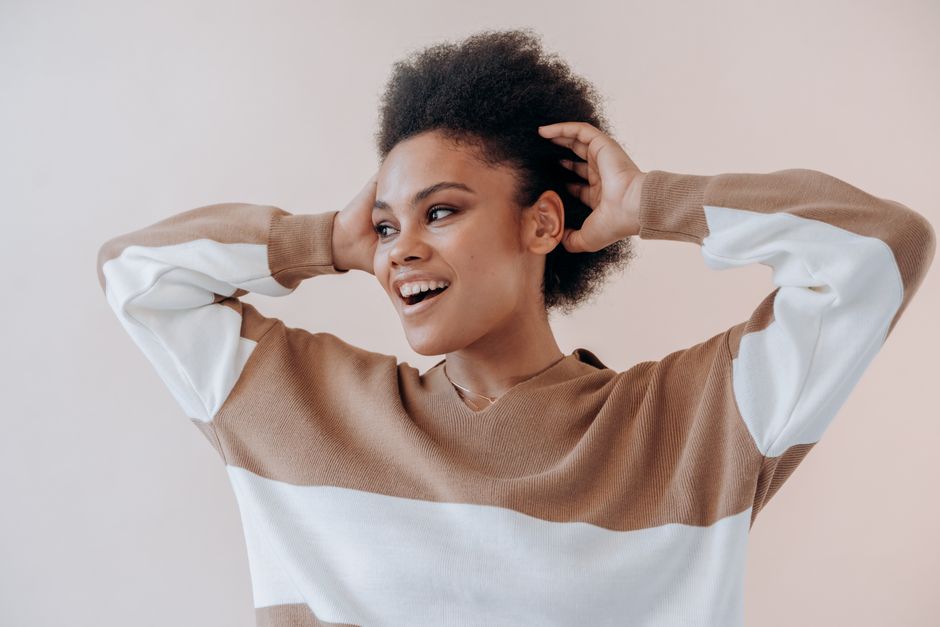 Happy woman with afro hair and striped sweater smiles indoors, enjoying a relaxed moment.