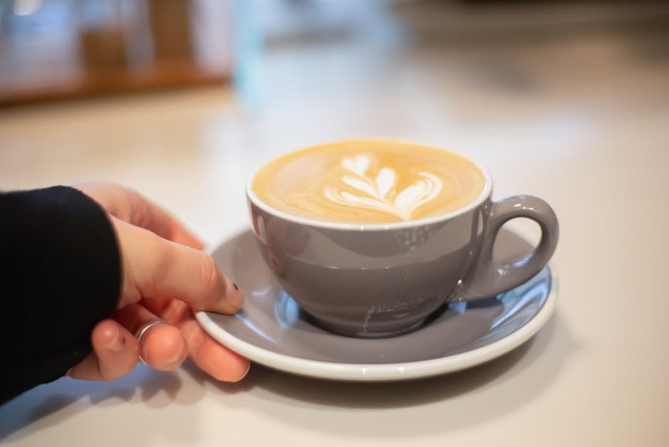 Hand holding a cappuccino with latte art in a cozy cafe setting. Perfect warm drink moment.