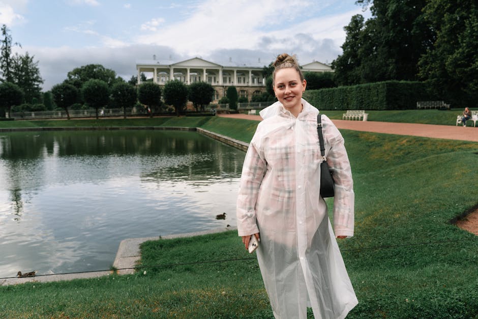 A woman in a white raincoat stands by a serene lake in a park, with a historic building in the background.