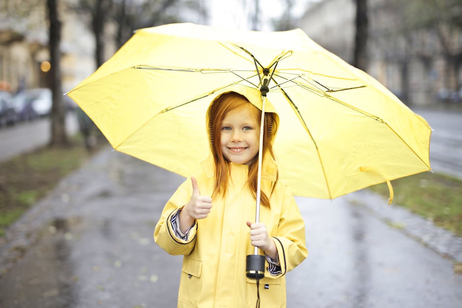 Young girl smiling under a yellow umbrella on a rainy street, showing a thumbs up.