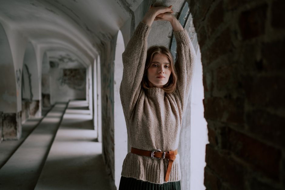 Woman in cozy sweater stretches by arched window in historic interior setting.
