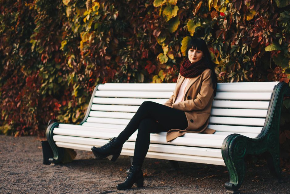 Woman in stylish autumn outfit sitting on a park bench with colorful leaves.