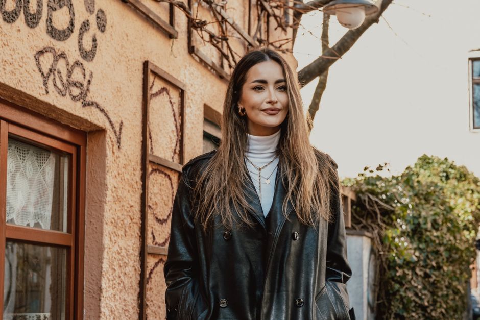 Fashionable woman in black leather coat standing against urban building backdrop.