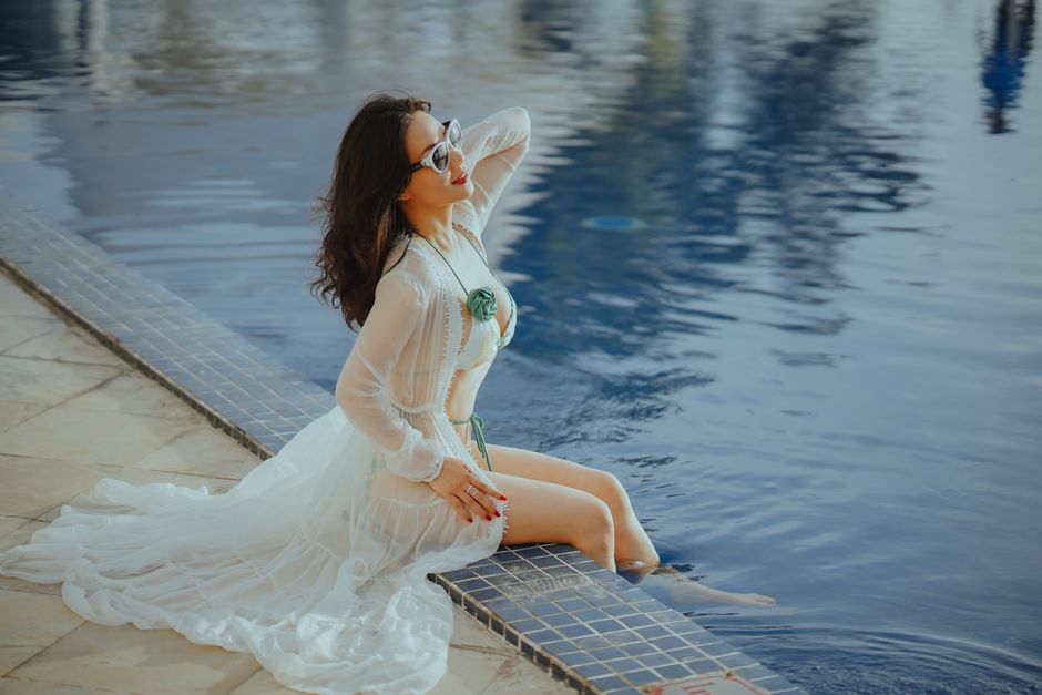 Elegant woman in a swimsuit with cover up, sitting by a poolside, wearing sunglasses.