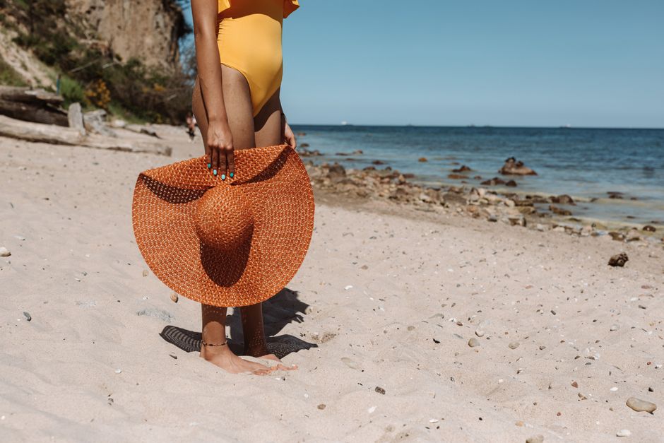 A woman stands on a sandy beach holding a large straw hat under the bright summer sun.