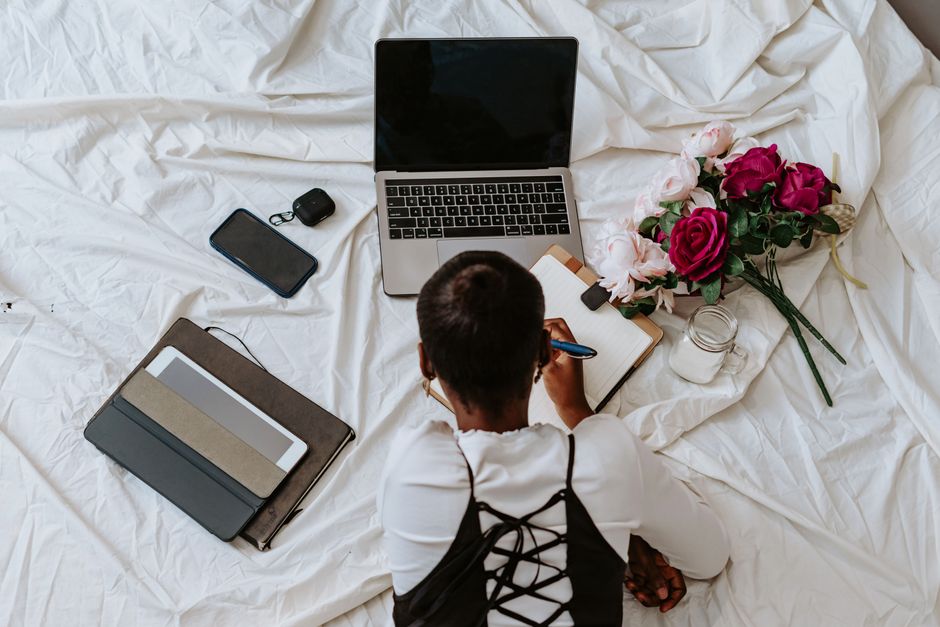A woman using her laptop and writing in a notebook surrounded by flowers and technology on a bed.