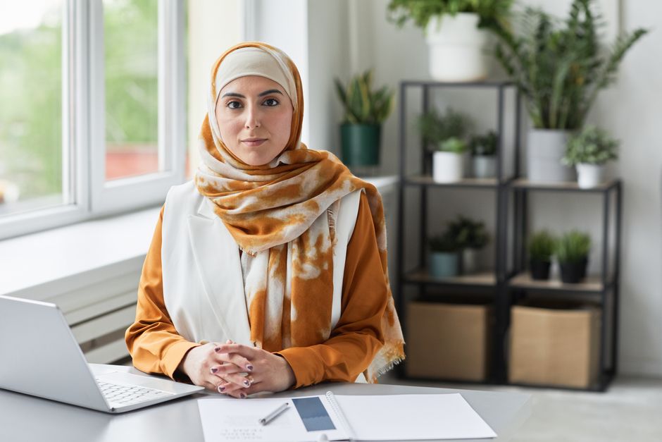 A focused Muslim woman in a headscarf working on a laptop in a modern office setting.