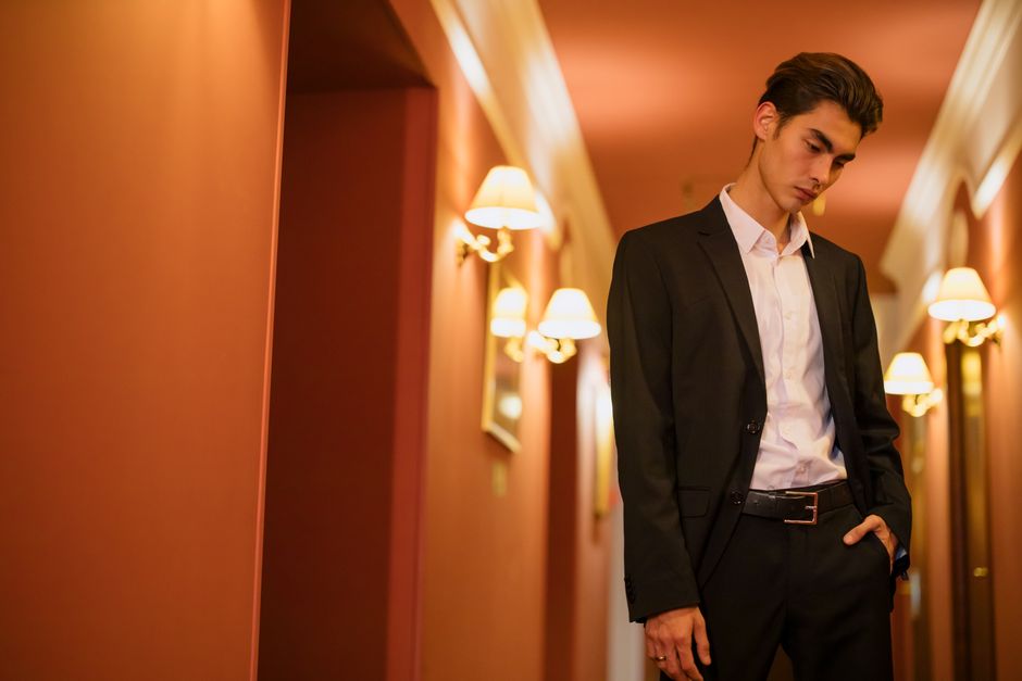 Young man in a black suit stands thoughtfully in an elegant hotel corridor.
