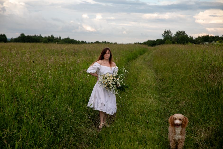 A woman in a white dress holding wildflowers walks with a dog in a lush green field.