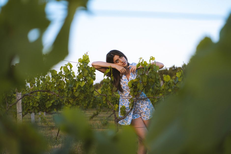 A woman in a summer dress poses playfully in a sunny vineyard.