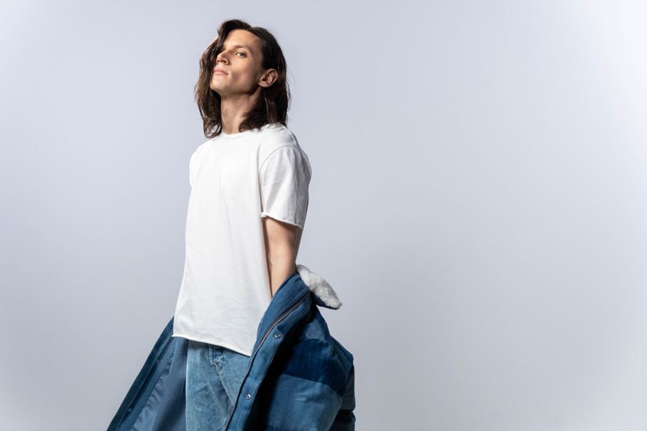 Man with long hair poses confidently wearing a white t-shirt and denim jacket against a white background.