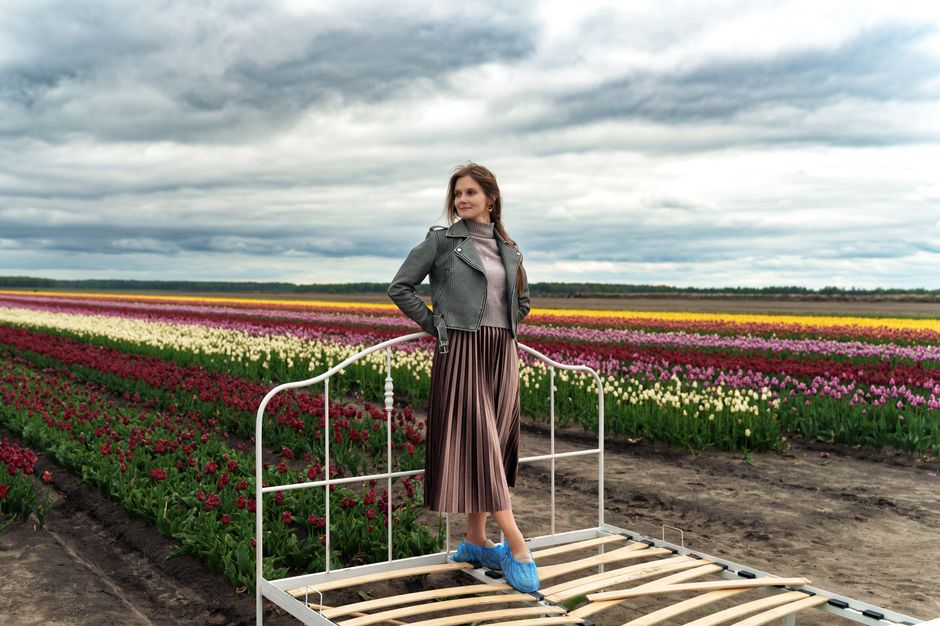 Young woman standing on bed frame in vibrant tulip field under cloudy sky.