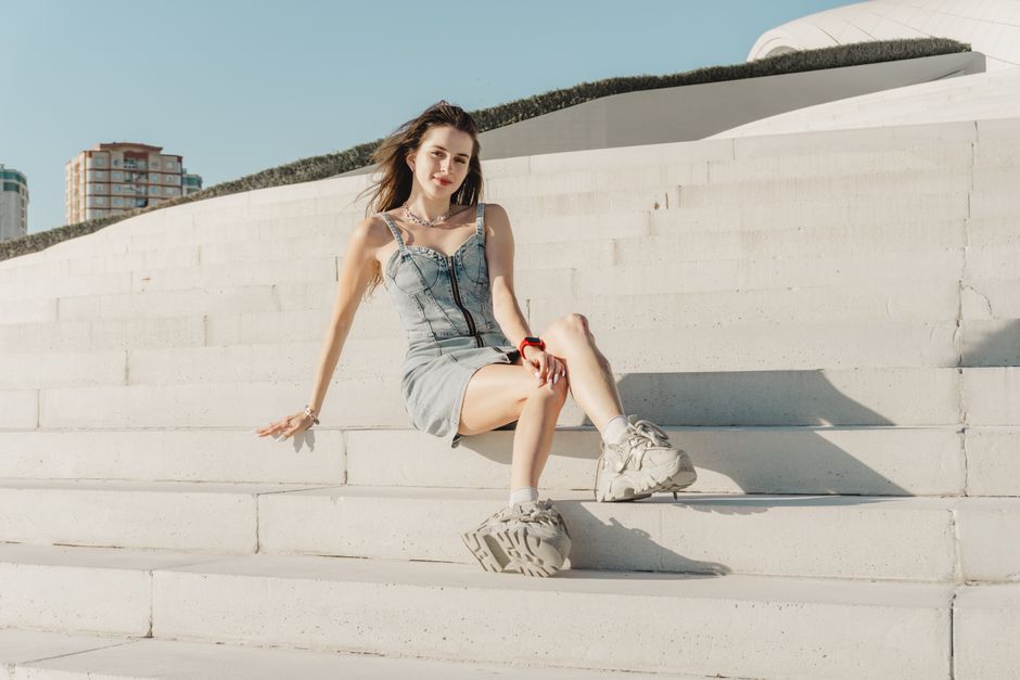 Young woman sitting on urban steps wearing a denim dress, enjoying a sunny day outdoors.