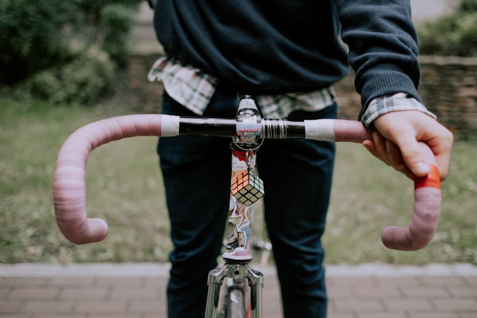 Faceless cyclist holds bicycle with pink handlebars and Rubik's Cube keychain in urban setting.