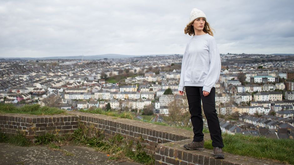 A woman standing on a hilltop overlooking Plymouth's cityscape, wearing a beanie and sweatshirt.