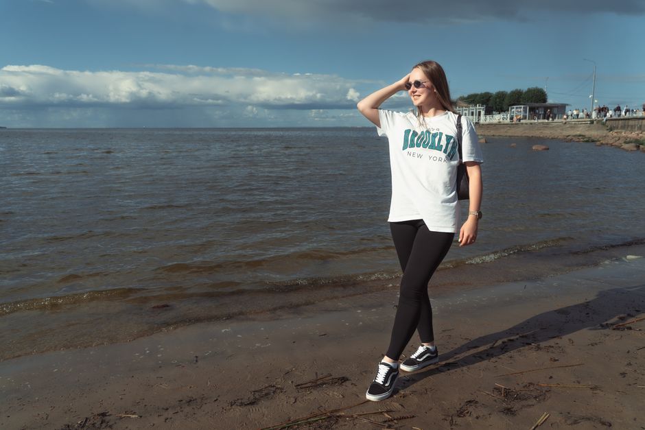 Young woman wearing sunglasses and casual outfit walking along the beach on a sunny day.