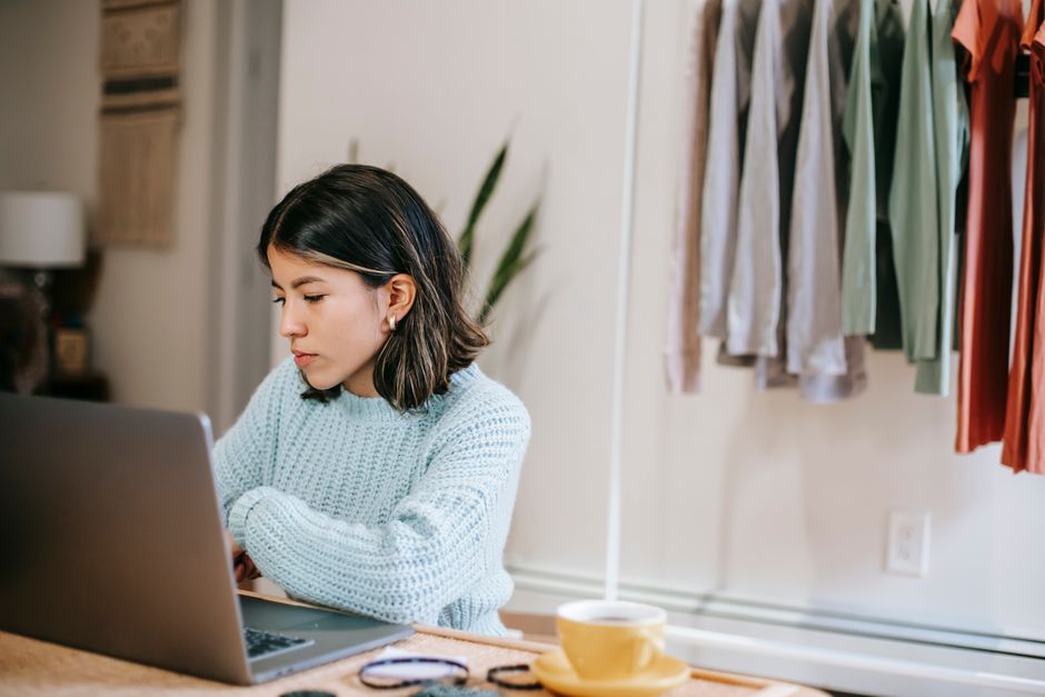 Concentrated young Latin American female freelancer in casual outfit browsing on laptop at table near cup with drink at light home with plant with green leaves near wardrobe with apparels